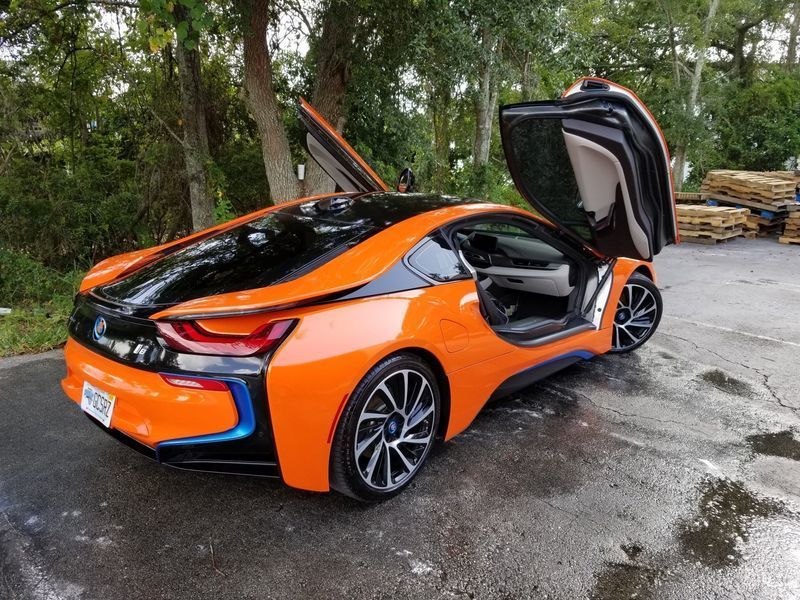 Orange and black sports car with butterfly doors open, parked on a wet road under trees.