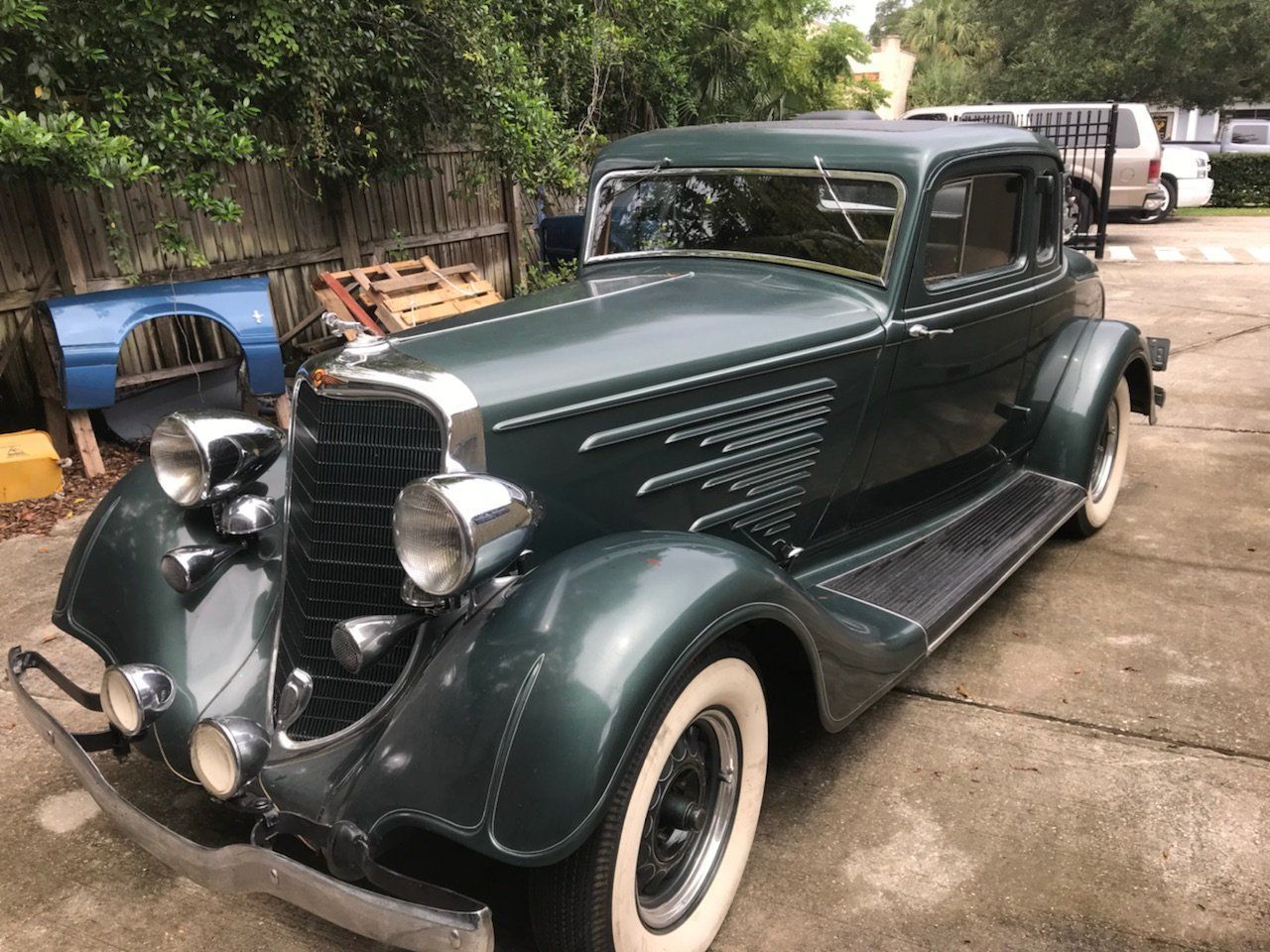 Vintage dark green classic car parked on a driveway, front-left view, with whitewall tires.