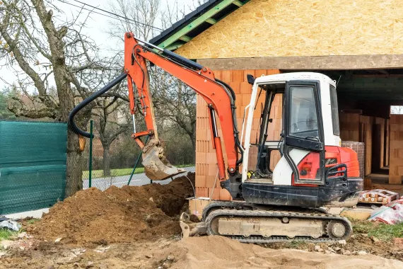 Orange and white excavator digging a trench next to a house under construction.