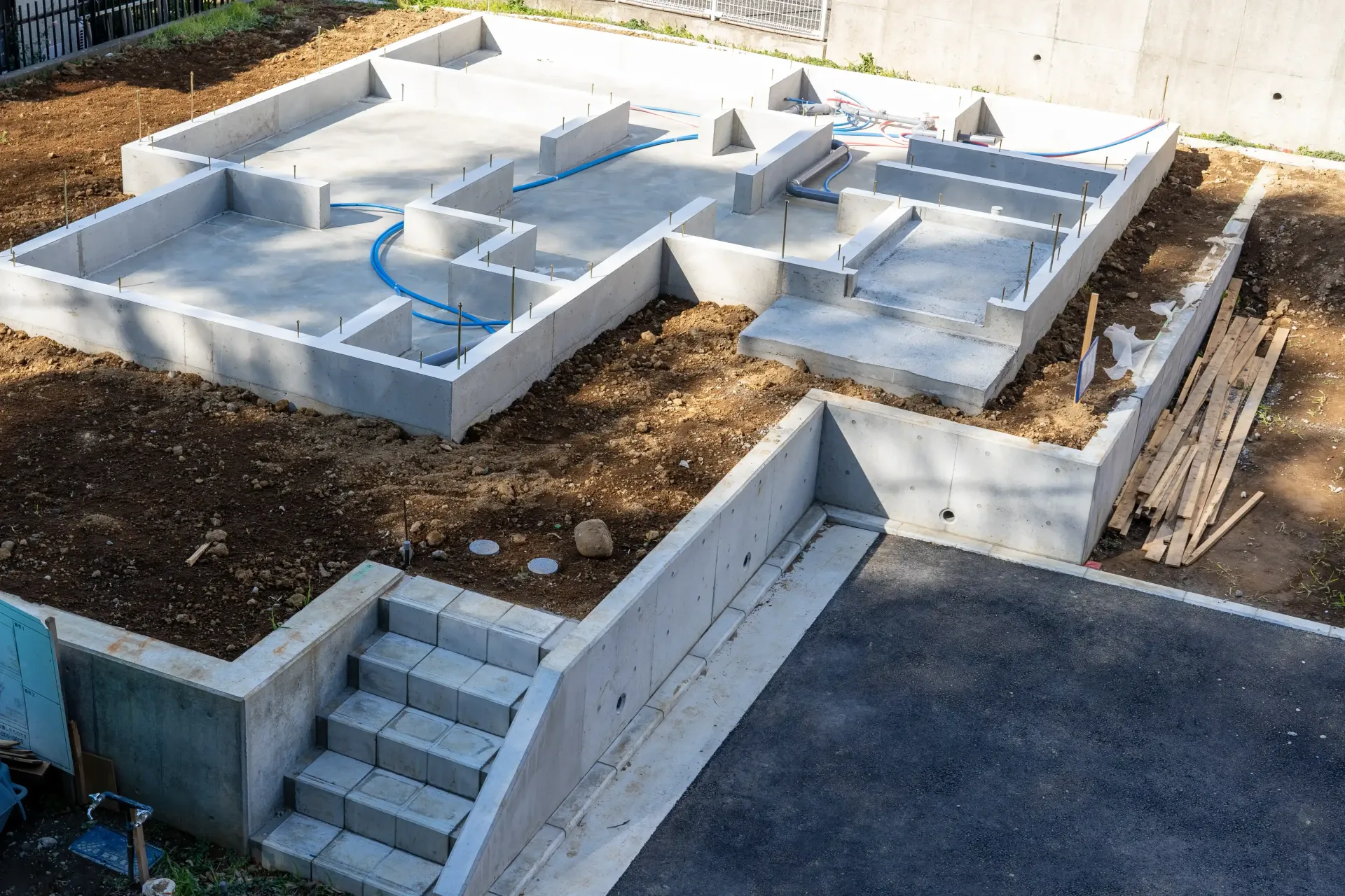 Concrete foundation of a building under construction, with steps leading down and blue pipes visible.