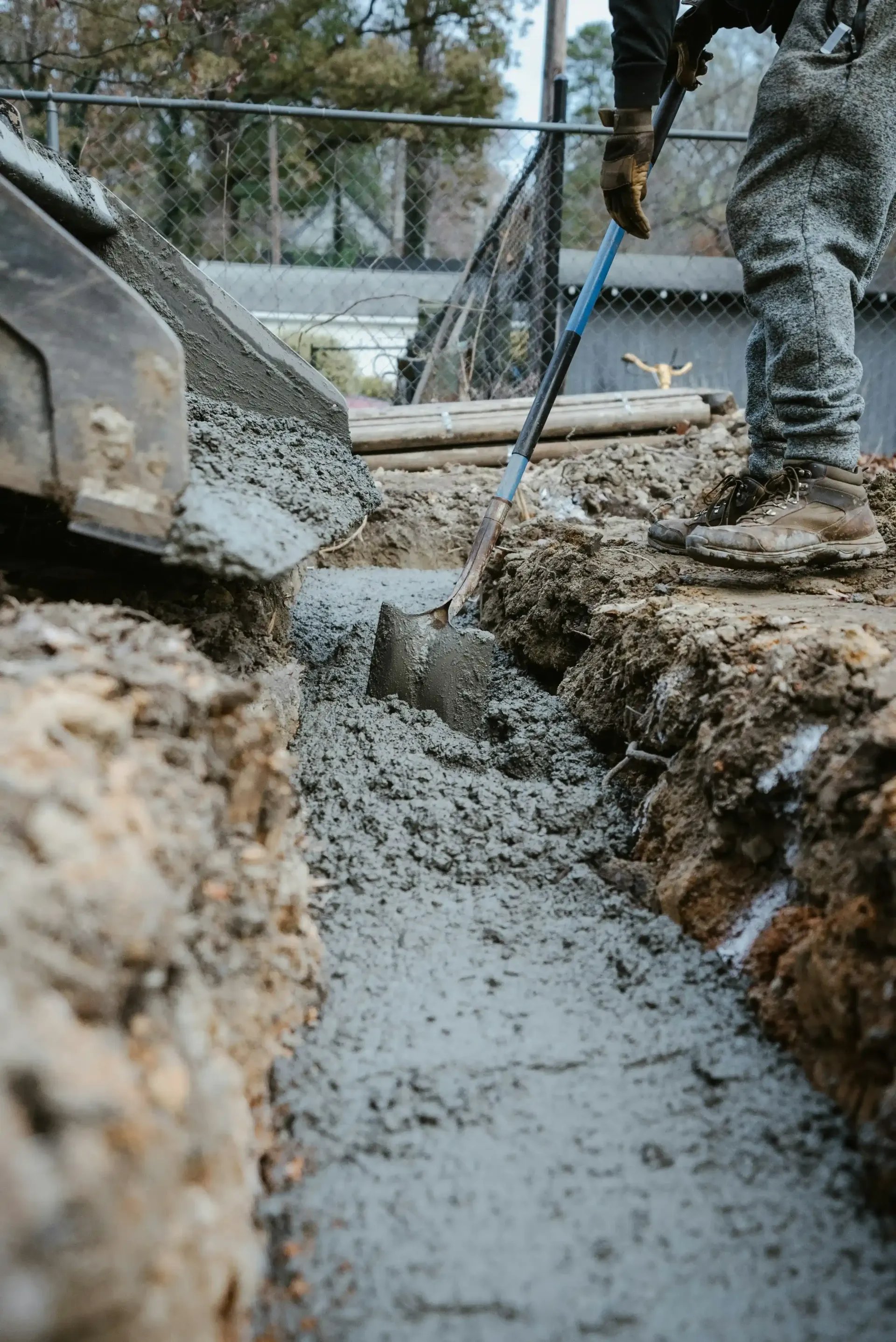 Construction worker leveling concrete in a narrow trench with a shovel; outdoor setting.