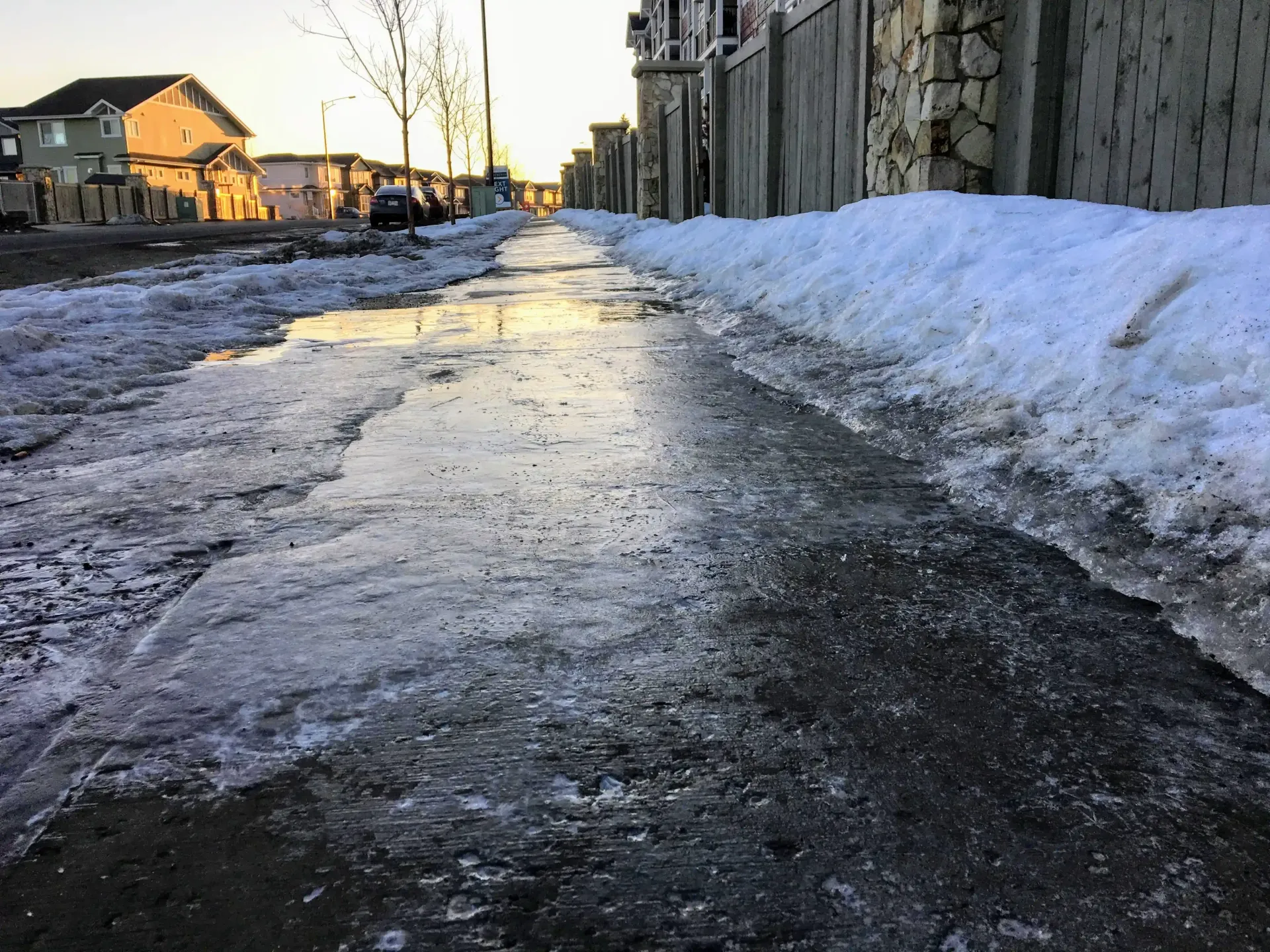 Icy concrete sidewalk with melting snow. Houses and fence in the background.
