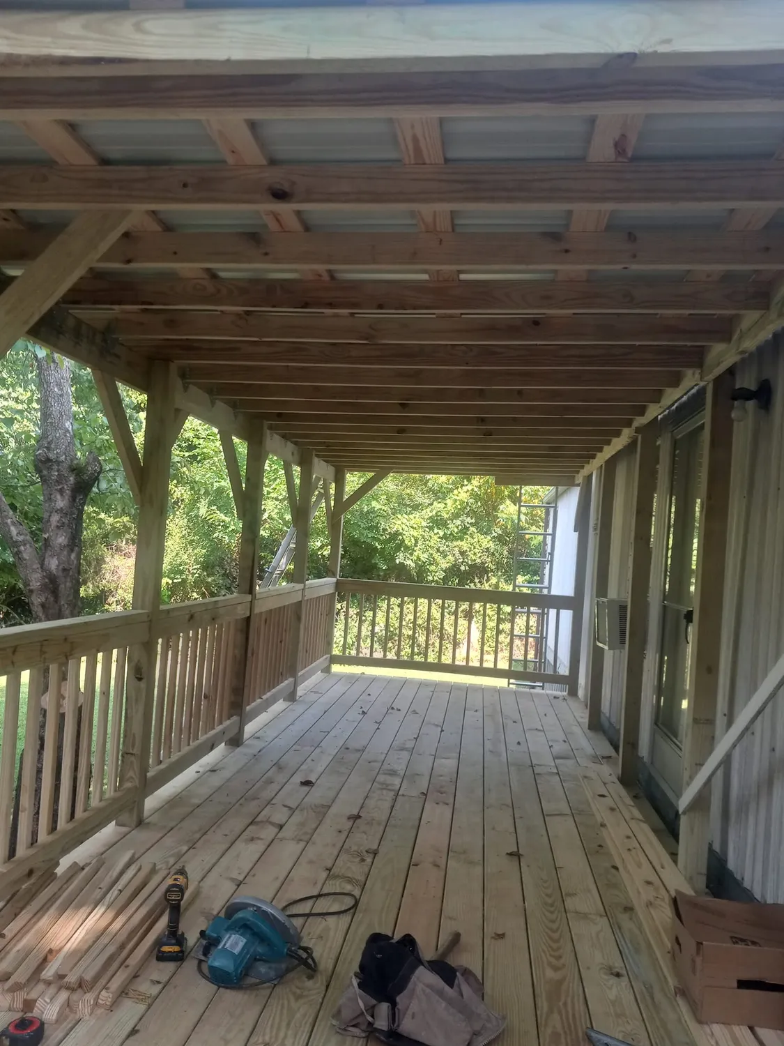 Wooden porch with railing, roof, and tools on the deck. Sunlight streams through trees in the background.