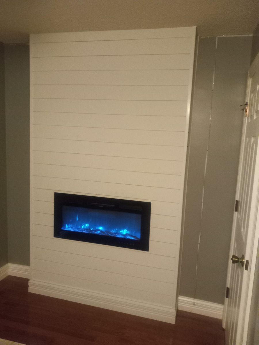 White shiplap accent wall with electric fireplace, blue flames, against gray walls, hardwood floor.