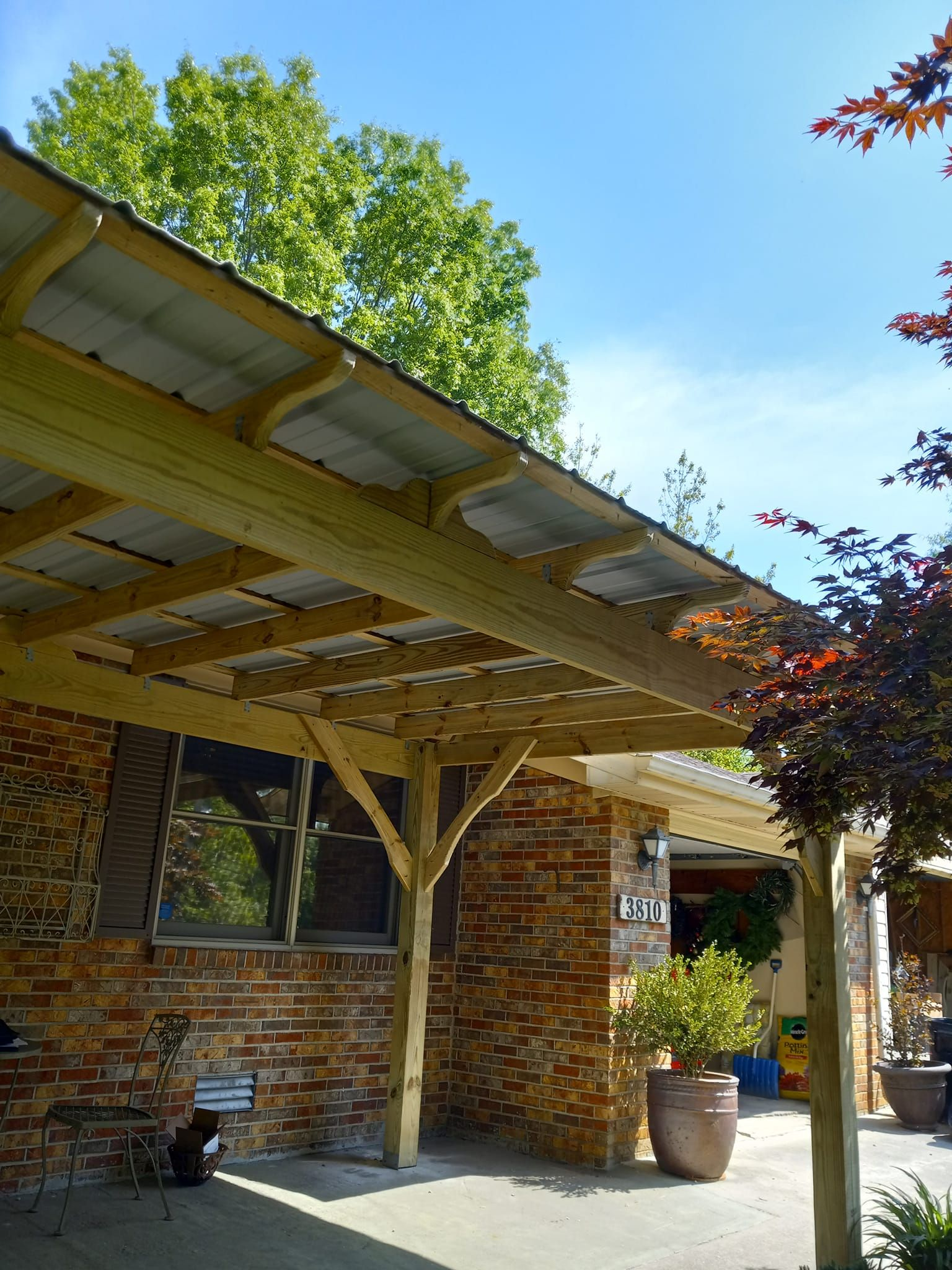 Wooden carport with corrugated metal roof attached to a brick building, sunny day.