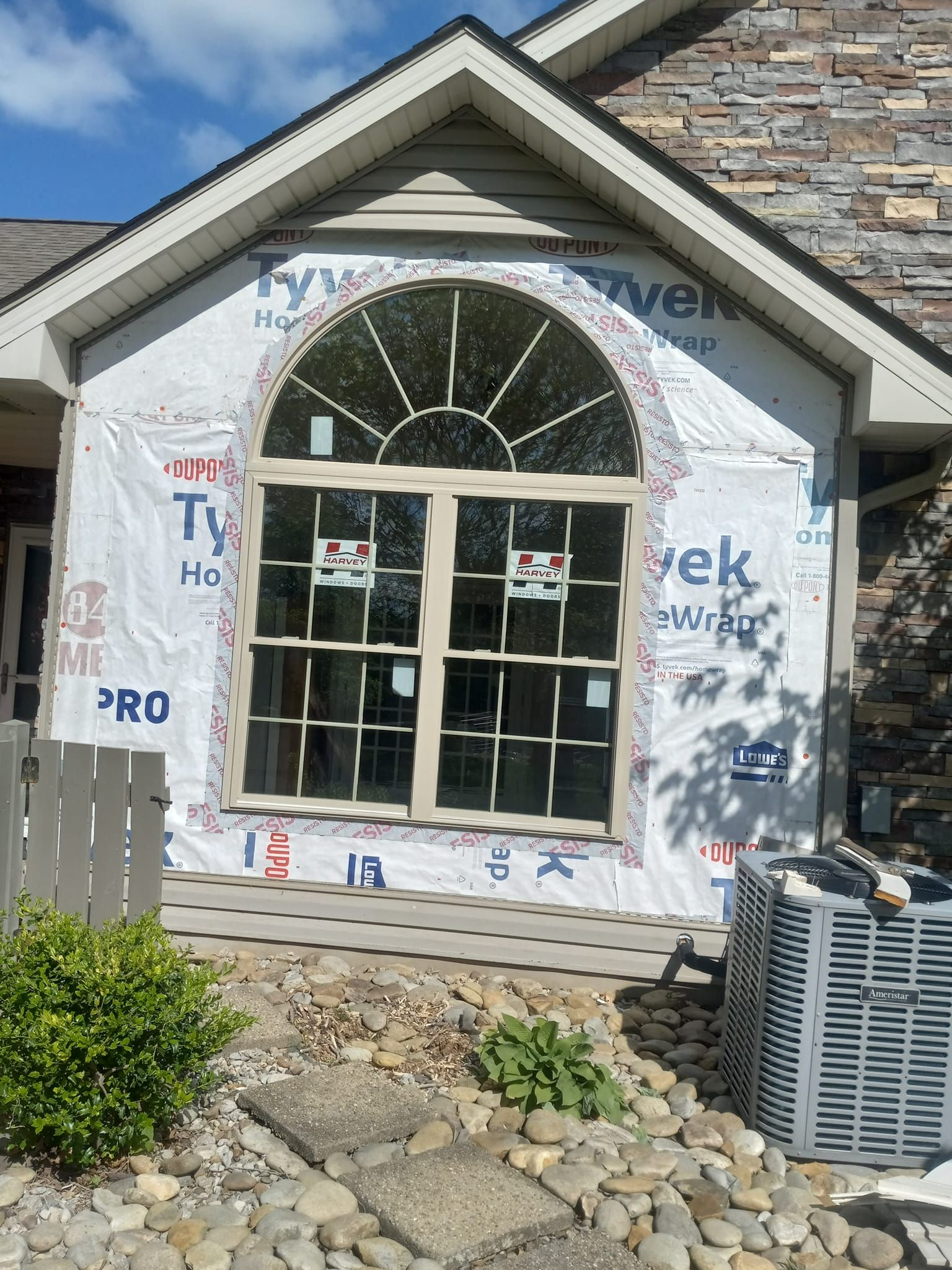 Exterior of a house with a large, arched window covered in Tyvek, an air conditioner, and landscaping.