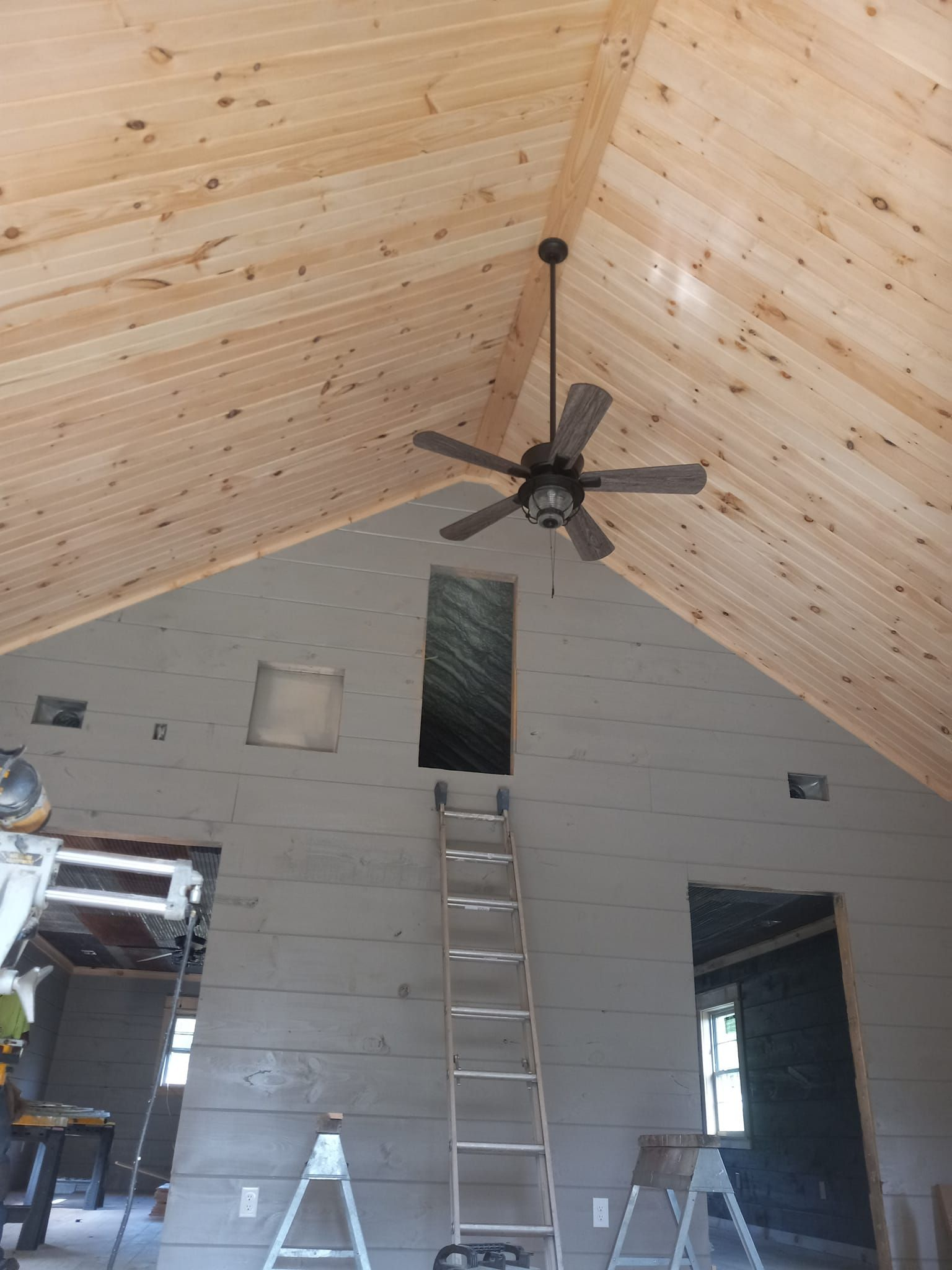 Interior of a cabin with a wooden ceiling, gray walls, a ladder, and a ceiling fan.