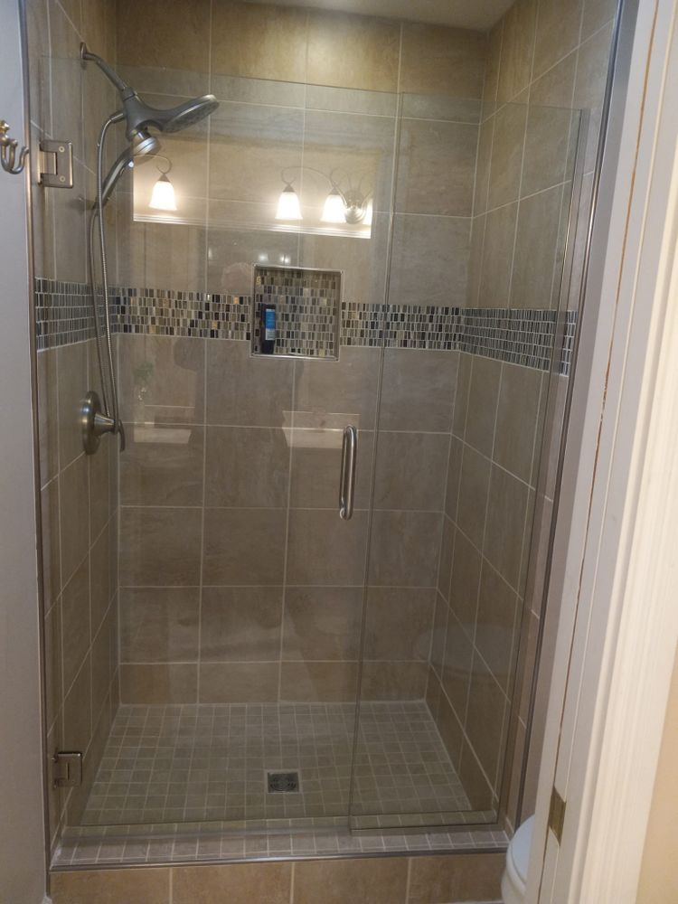 Tiled shower with glass door, showerhead, and shelf. Beige tiles with blue and brown mosaic accent.