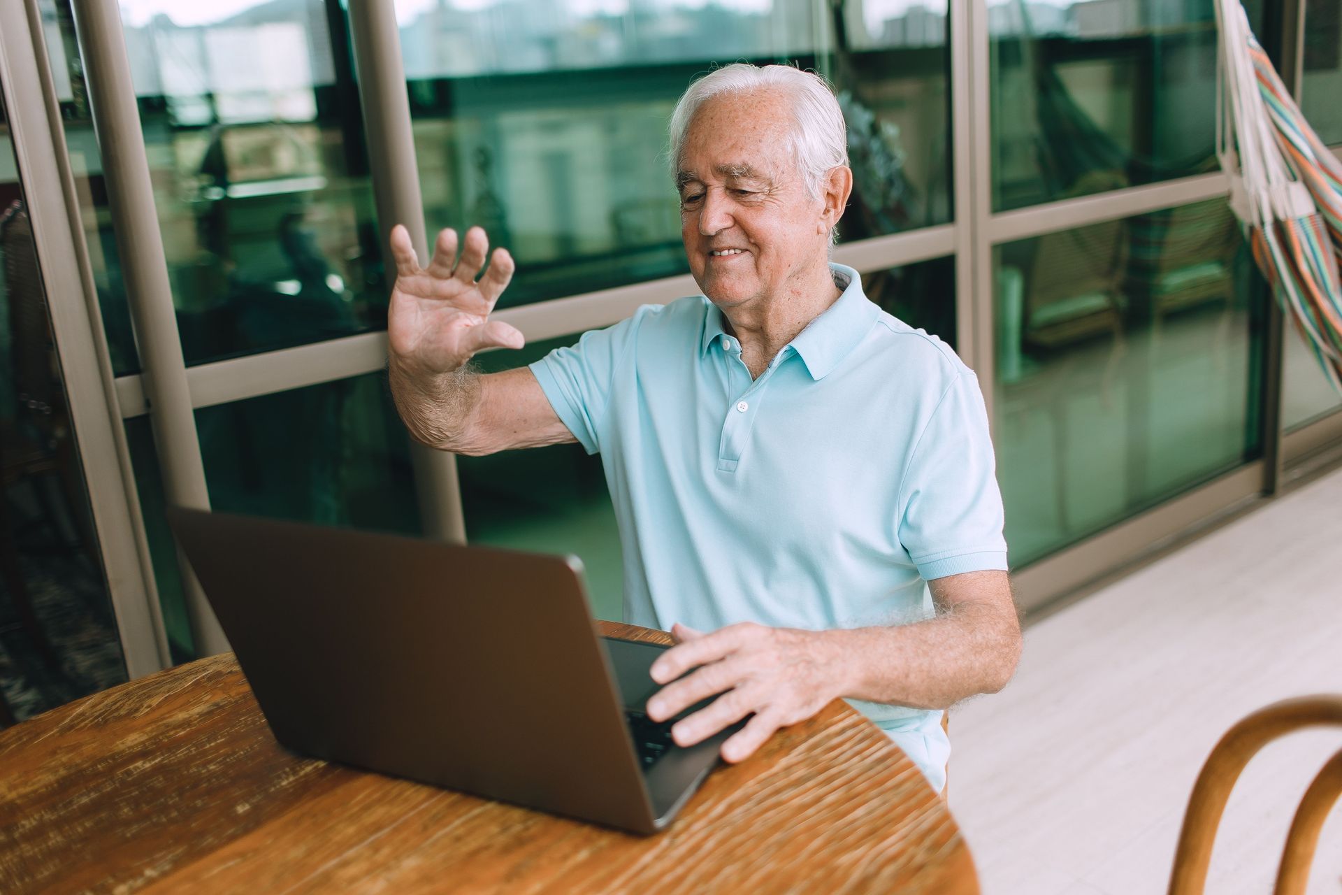 Smiling man in a light blue shirt using a laptop at a wooden table, waving in a bright modern hallway