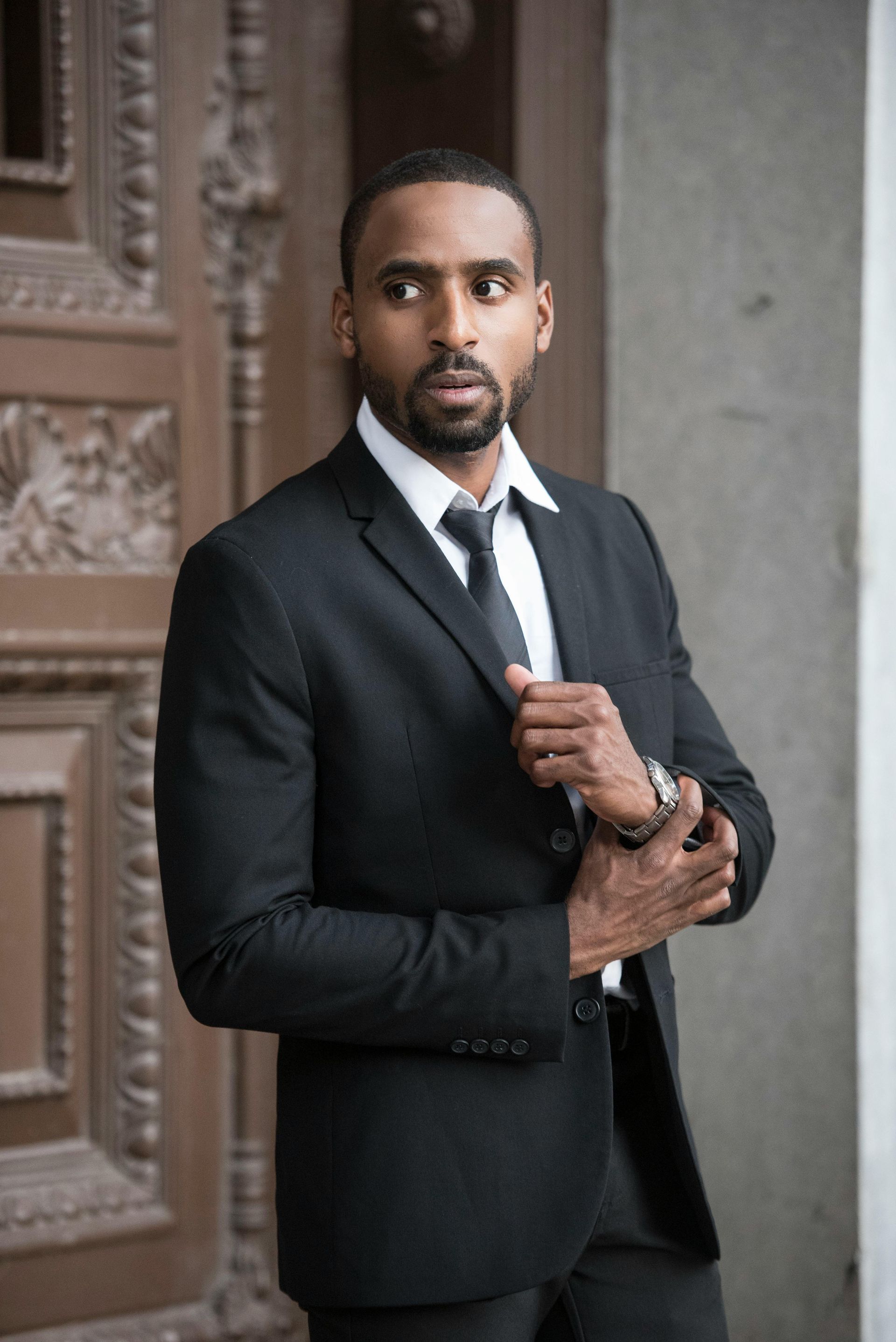 Man in black suit, adjusting jacket, standing near a wooden door.