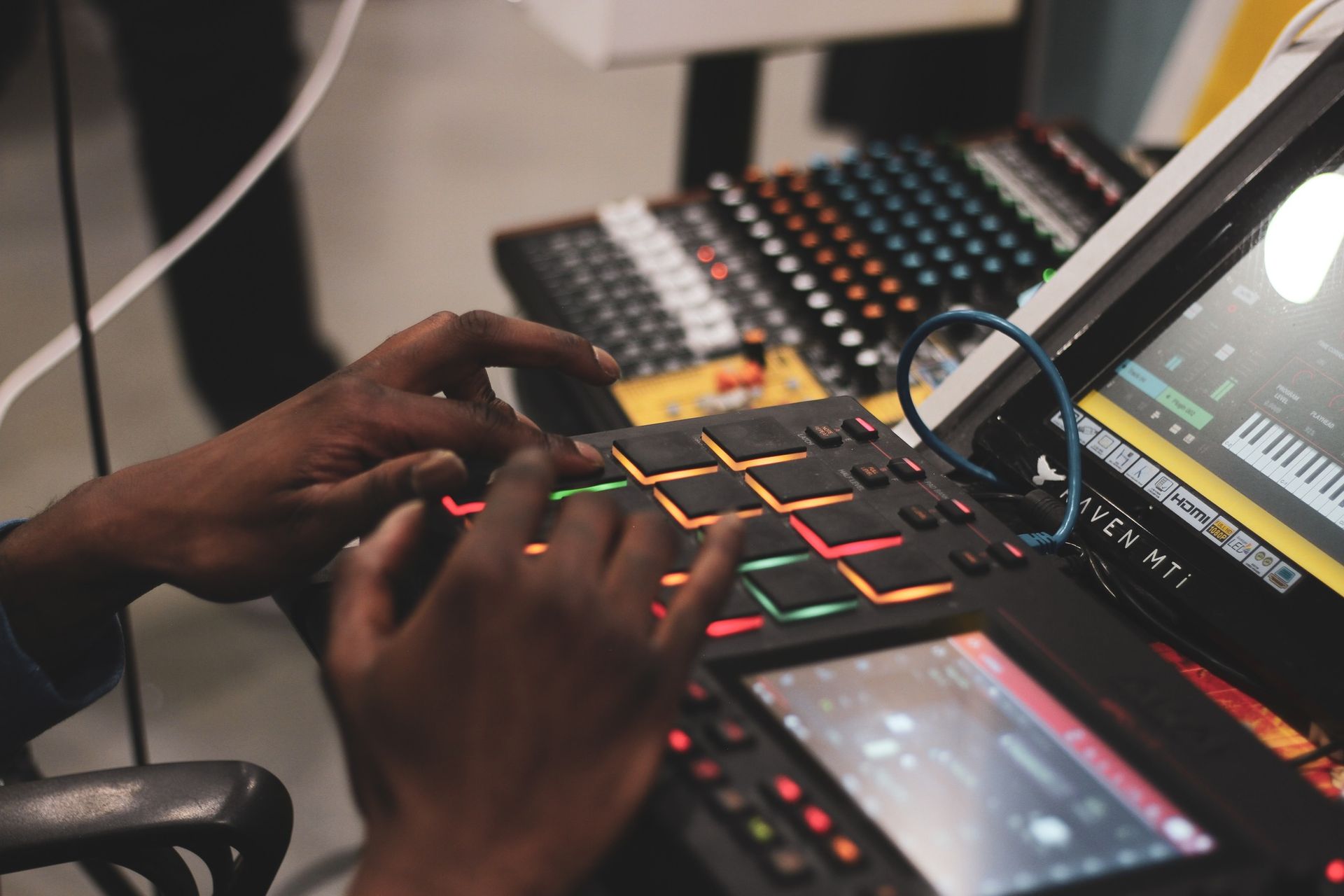 A person's hands playing an electronic drum pad, with a mixing board and a laptop in the background.