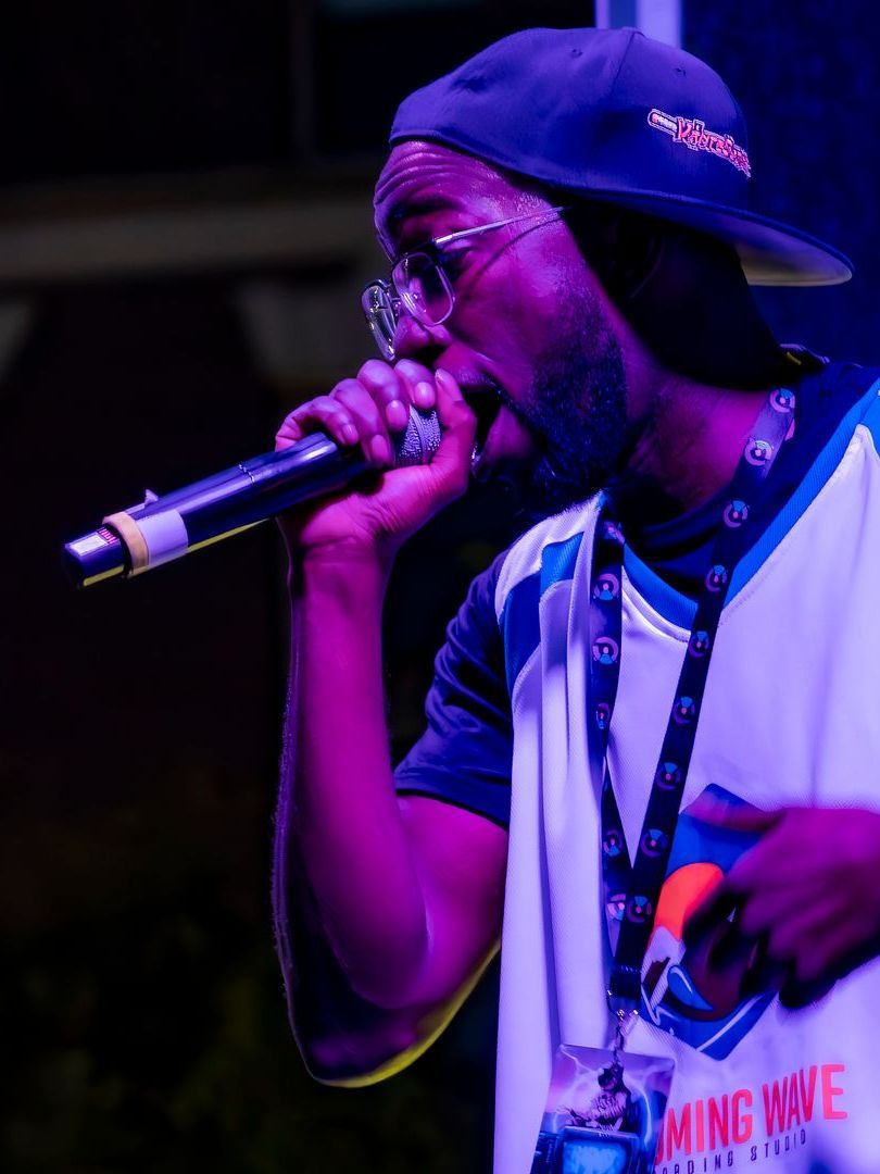 Man rapping into a microphone, wearing a baseball cap, glasses, and a jersey, lit by purple light.