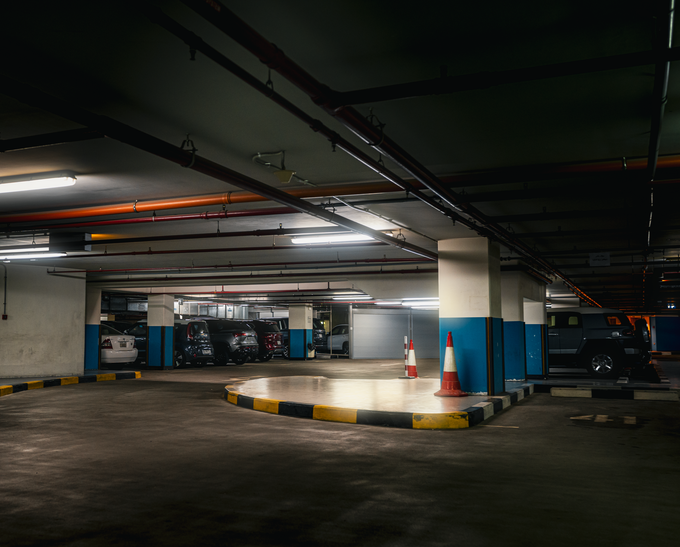 Autorimessa sotterranea poco illuminata, con pilastri di cemento, cordolo dipinto e auto parcheggiate.