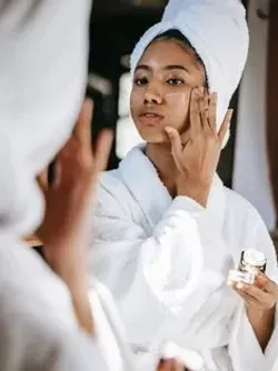 A woman with a towel wrapped around her head is looking at her face in the mirror