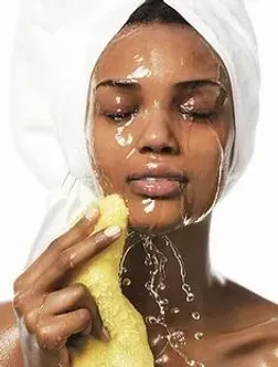 A woman with a towel wrapped around her head is washing her face with a sponge