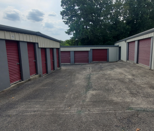 Storage units with red doors and gray siding. Paved lot. Green trees in the background. Overcast sky.