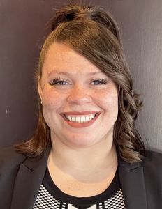 Woman smiling, wearing a black blazer and patterned top. Her hair is partially pulled up.