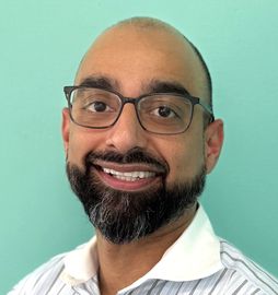Man wearing glasses, smiling with a full beard, against a light blue backdrop.
