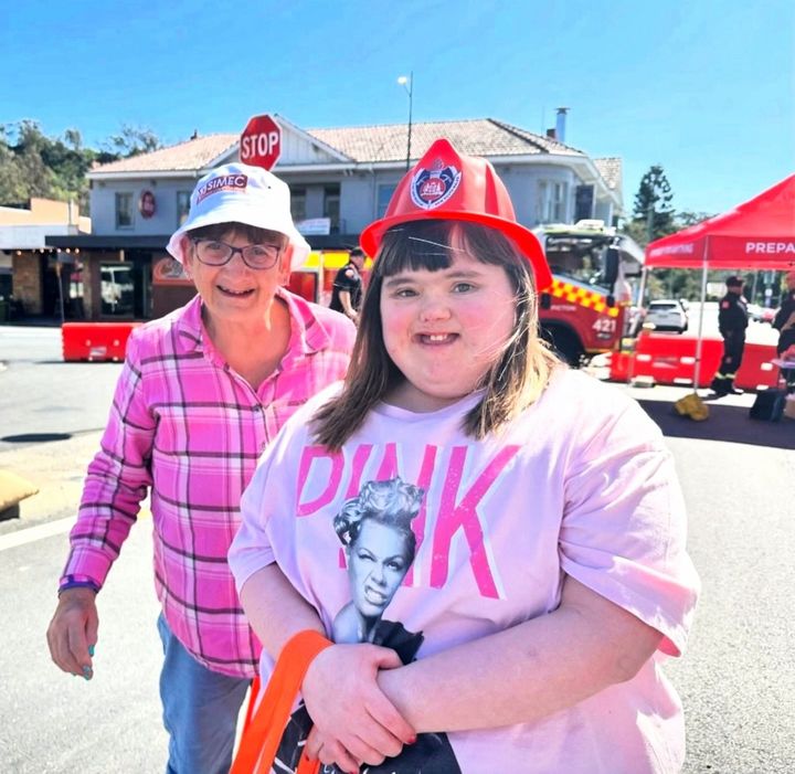 Two people in fire hats standing on a sunny street with emergency vehicles in the background