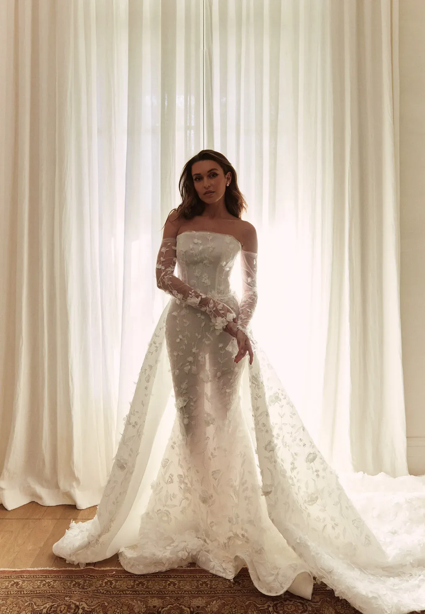 Woman wearing a white, off-the-shoulder wedding gown with long train, standing in front of a window with white curtains.