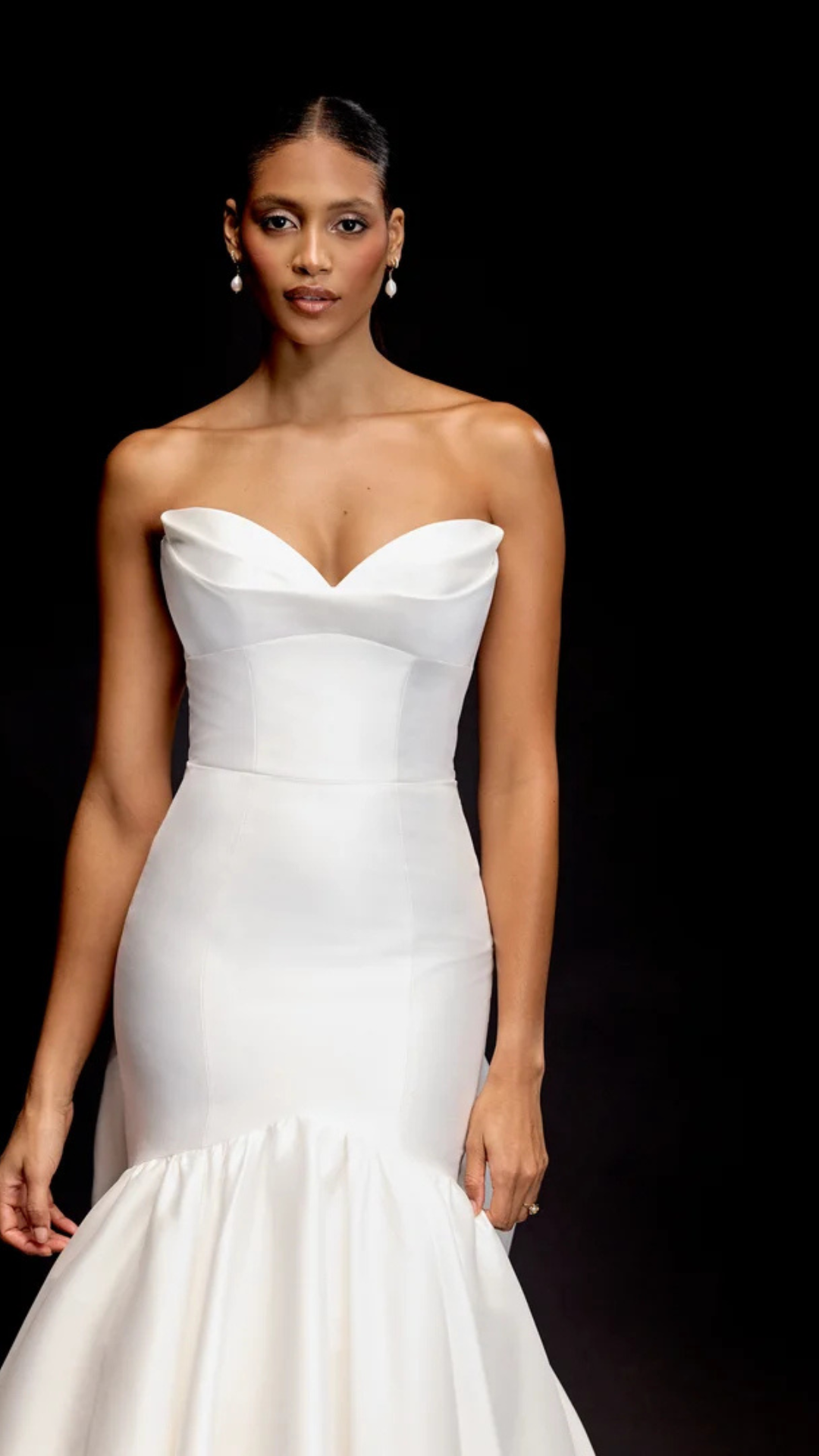 Woman in white strapless wedding gown, fishtail skirt, standing against black backdrop.