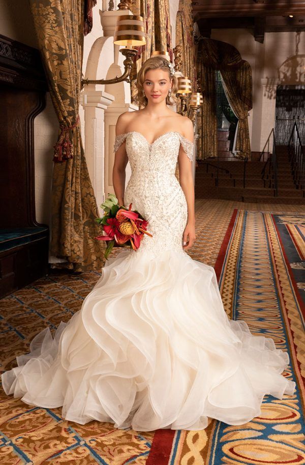 Woman in a white off-the-shoulder wedding dress with a ruffled skirt, holding flowers, posing in an ornate interior.