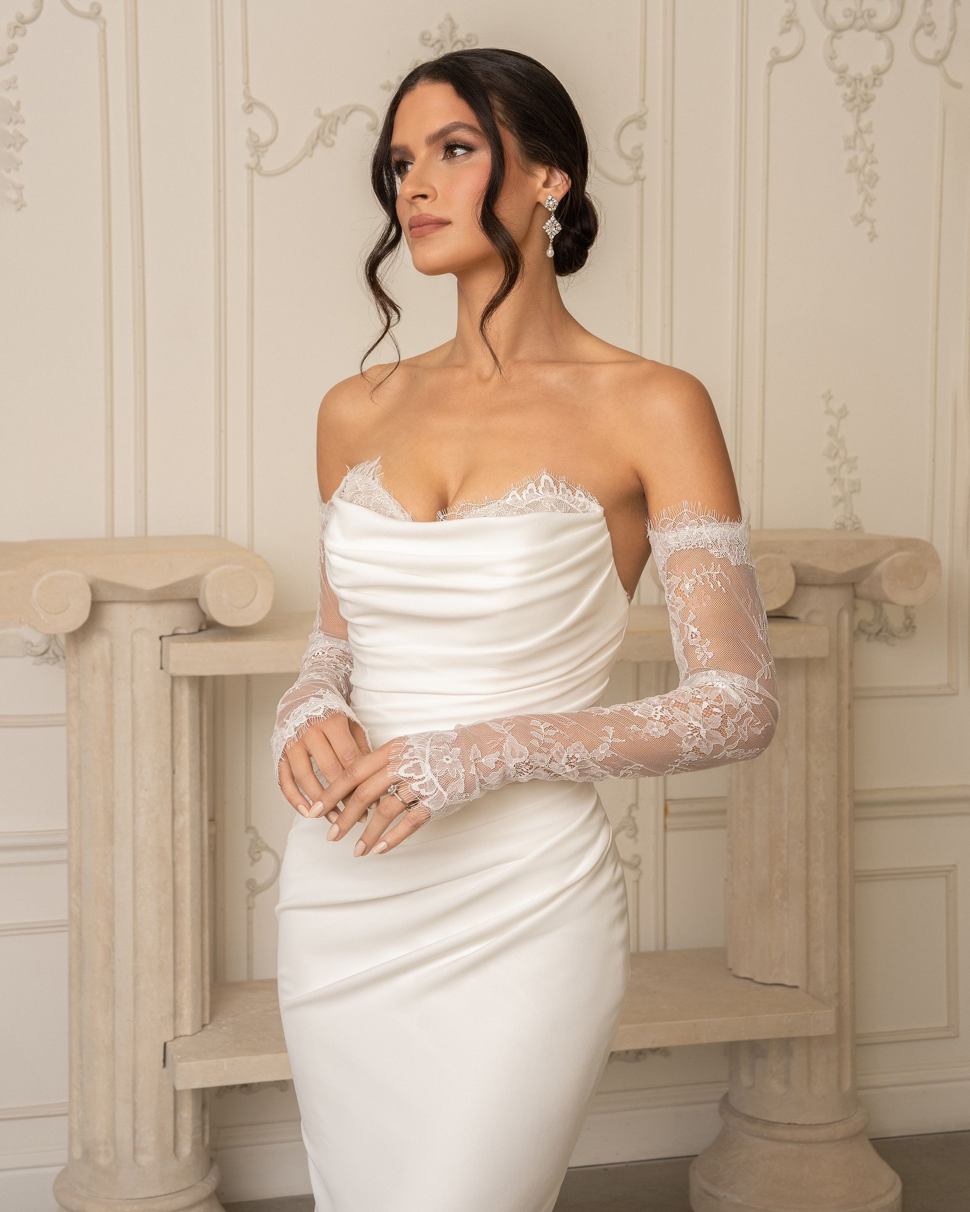 Woman in white wedding dress with lace sleeves, standing indoors.