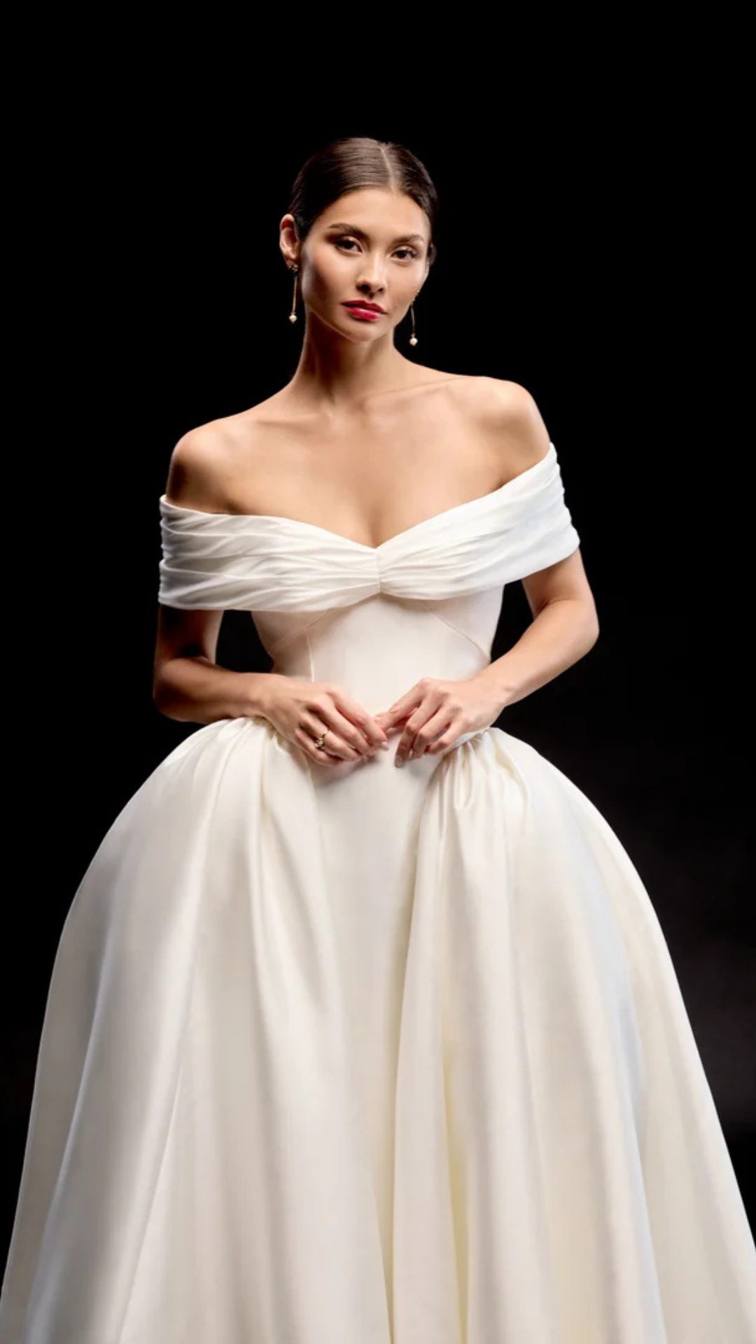 Woman in a white off-the-shoulder ball gown with a gathered bodice, standing against a black background.