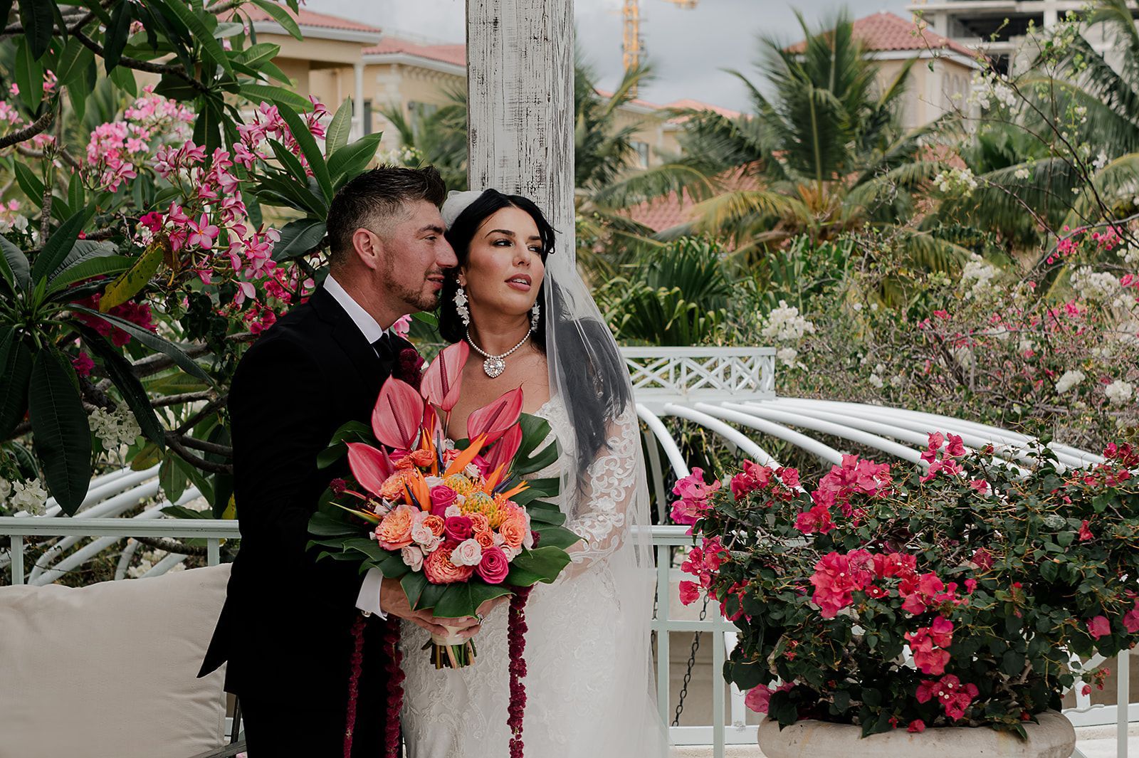 Wedding couple on a balcony with tropical flowers; the bride holds a colorful bouquet, and the groom embraces her, looking out at a vibrant garden.