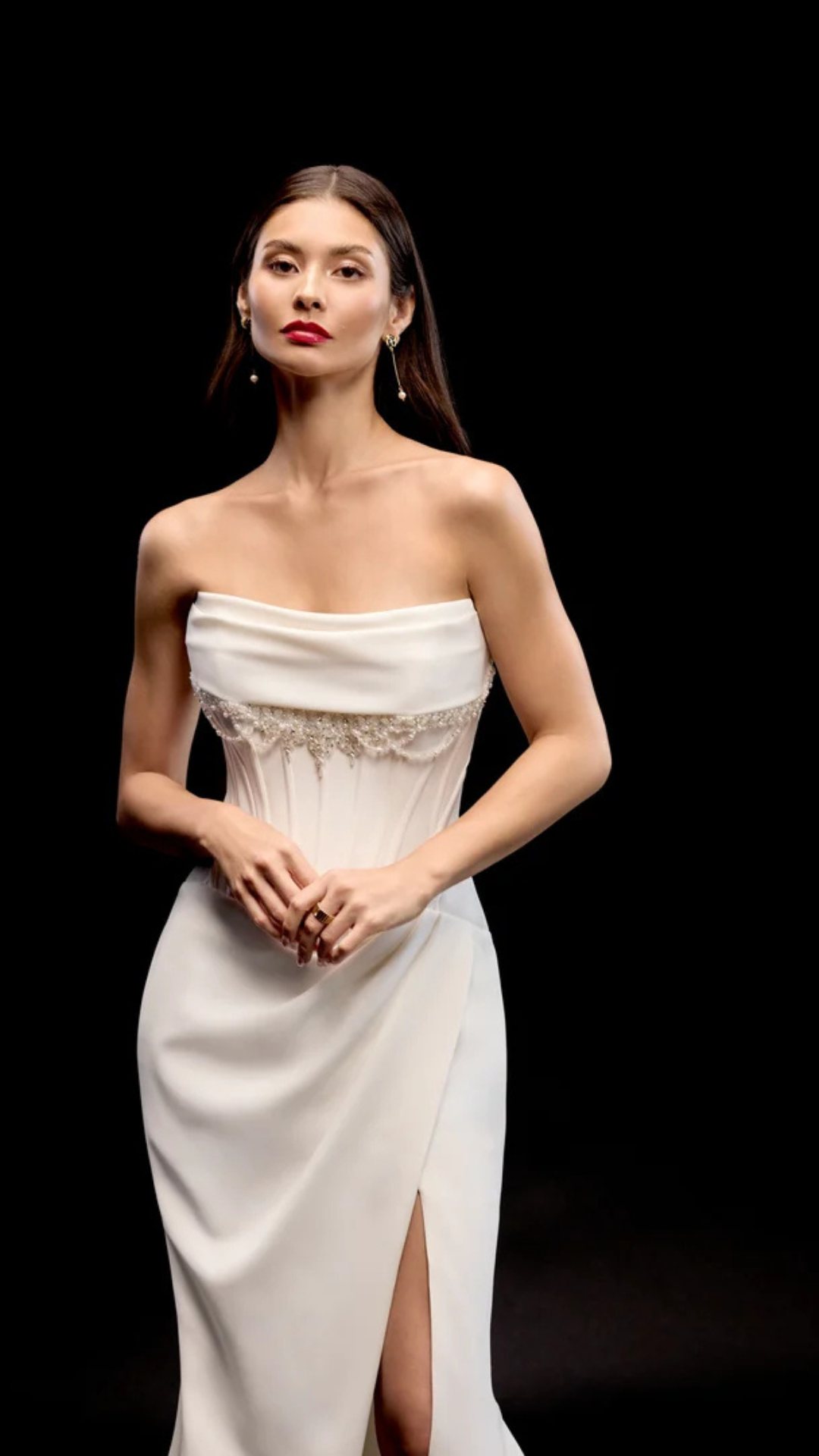 Woman in white strapless dress, dark background, looking forward, hands clasped.