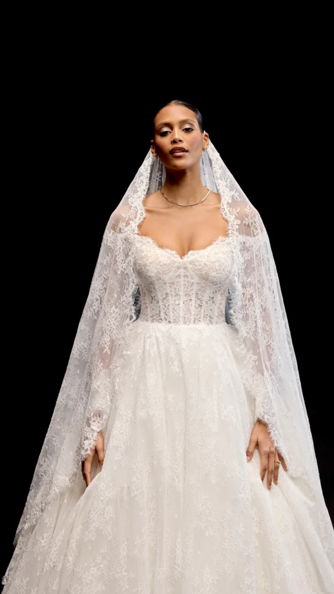Woman in a white lace wedding dress and veil, standing against a black background.