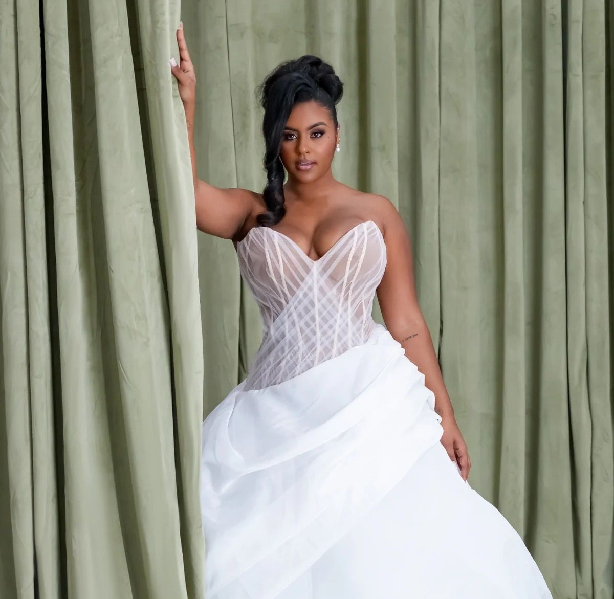A woman in a white wedding dress is standing in front of a grey curtain.