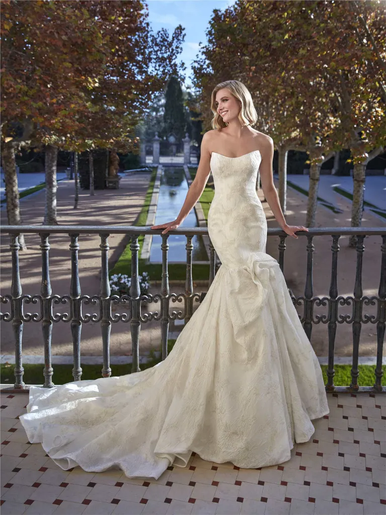 Woman in a strapless white wedding dress with a long train, standing on a balcony.