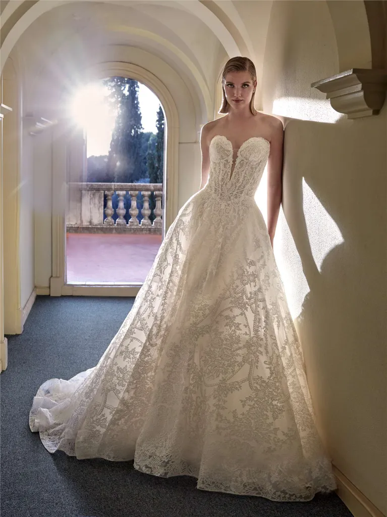 Woman in strapless white wedding dress leans against a wall in a hallway with a bright window.