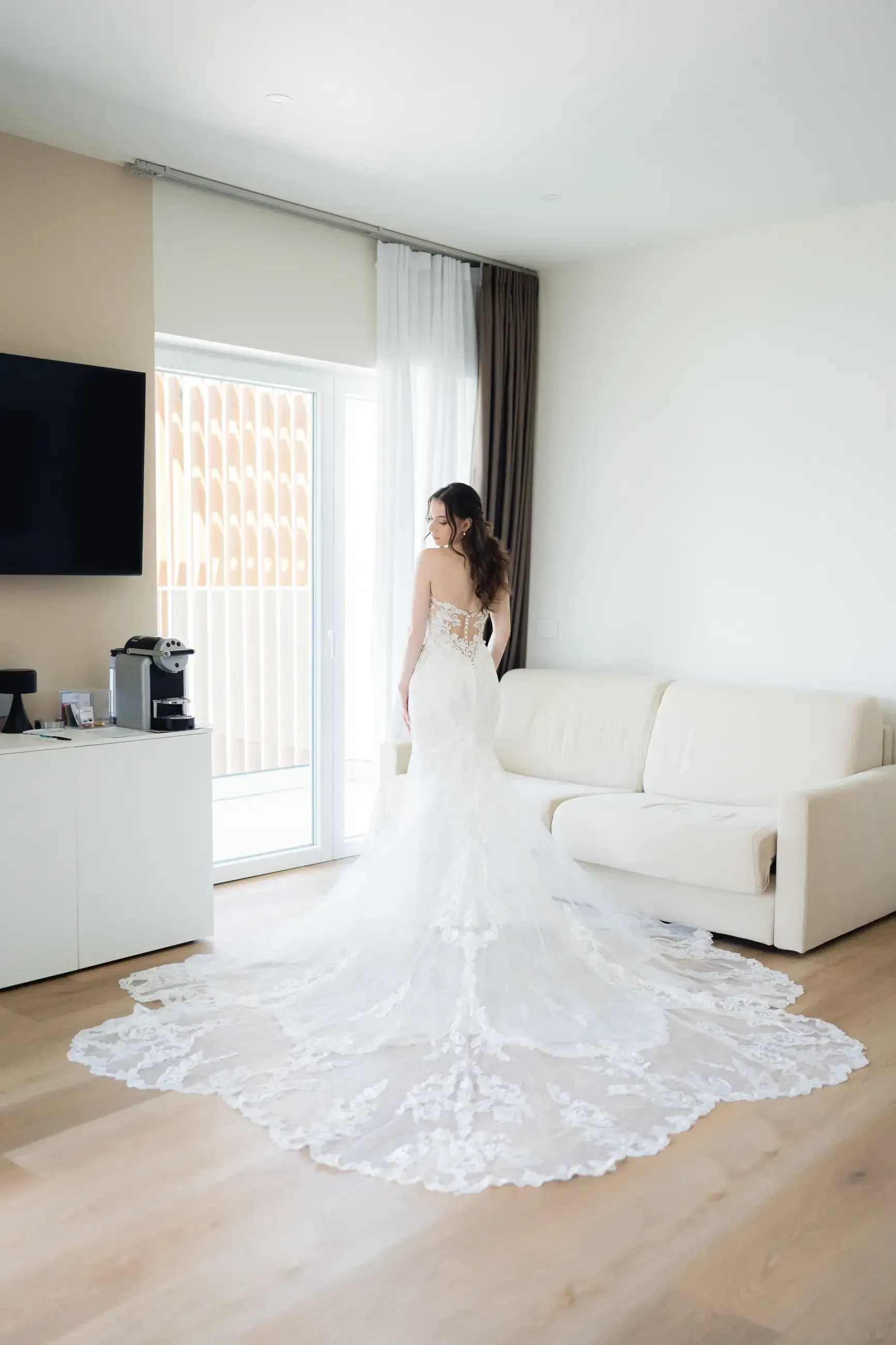 A woman in a wedding dress is standing in a living room looking out of a window.