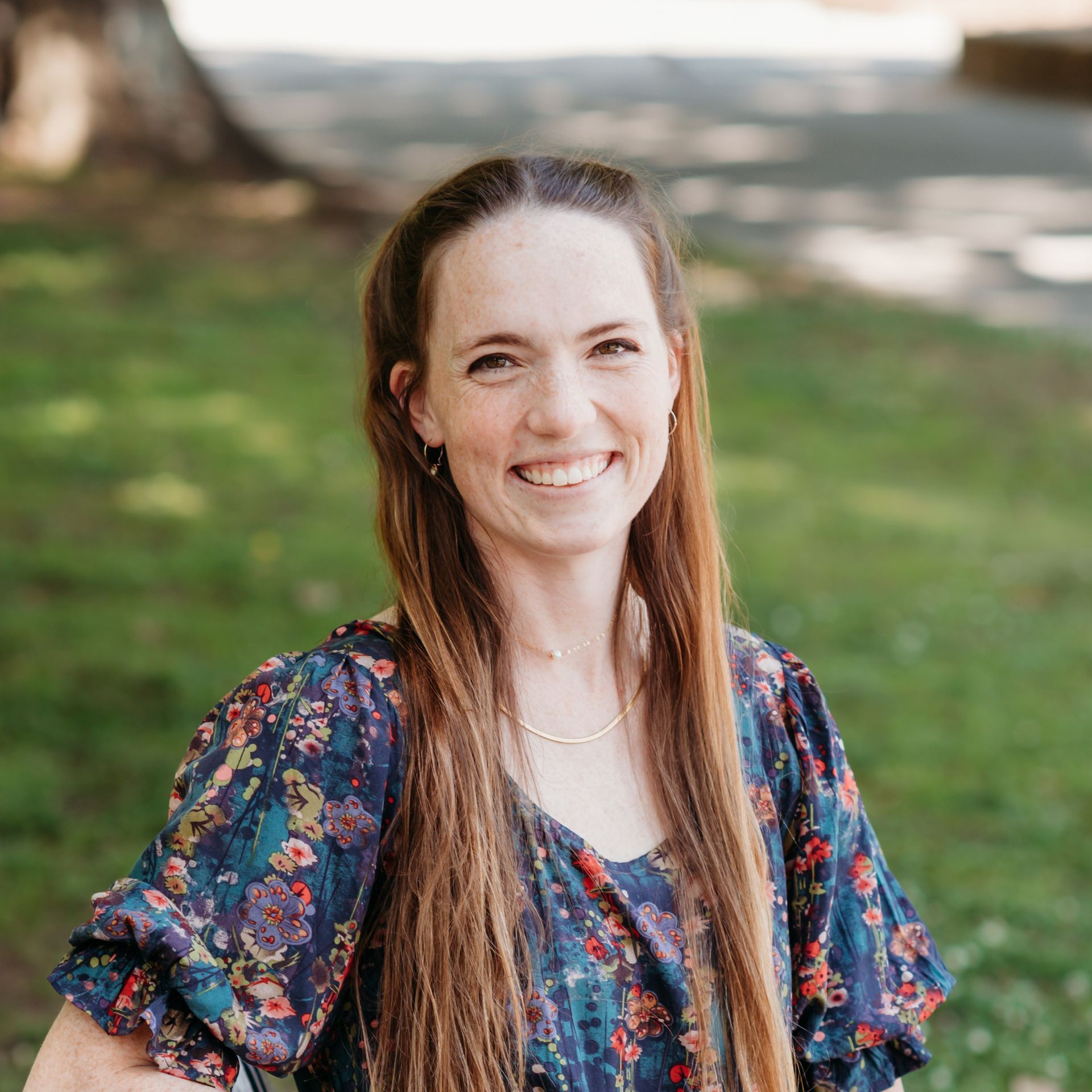 A woman with long hair is smiling for the camera while standing in the grass.