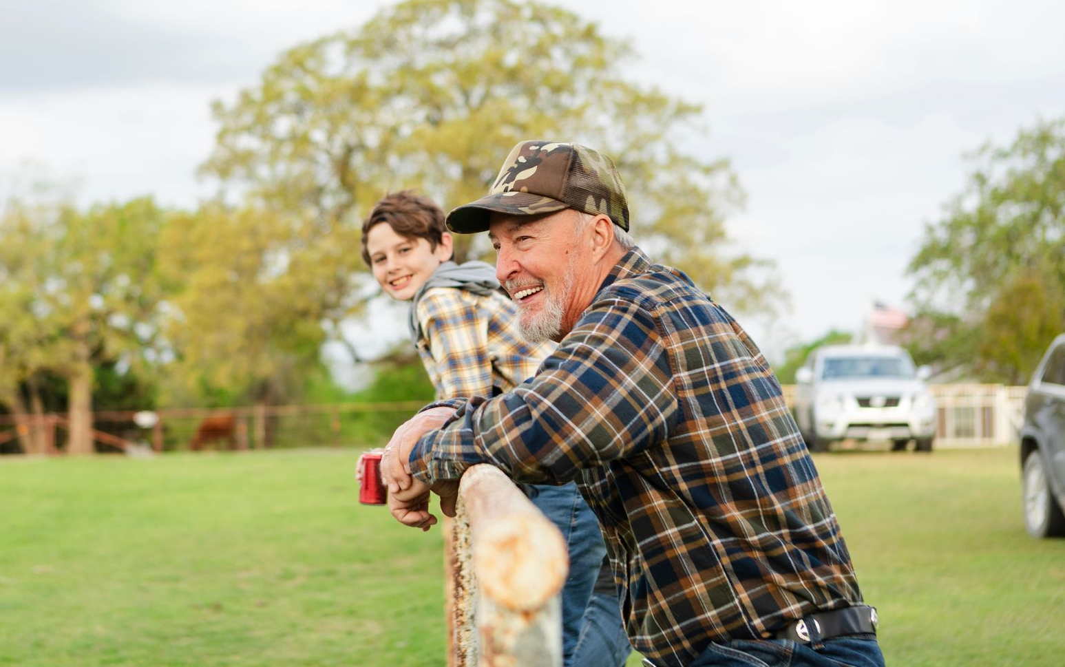 Two people leaning on a fence in a grassy field, smiling outdoors on a sunny day.