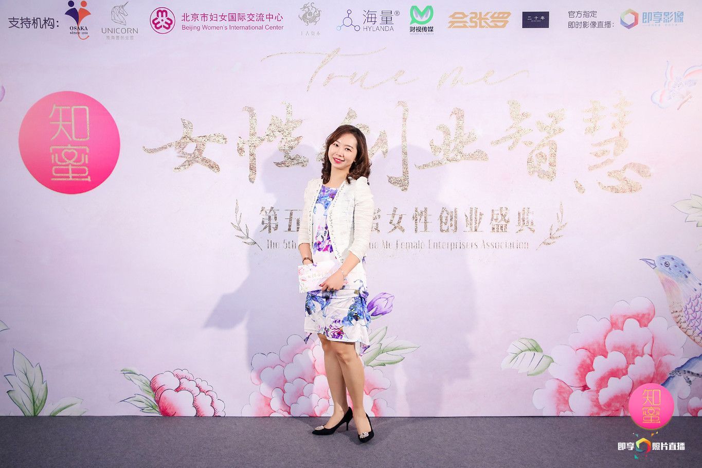 穿著碎花洋裝的女子在印有中文文字和標誌的正式活動背板前擺姿勢。