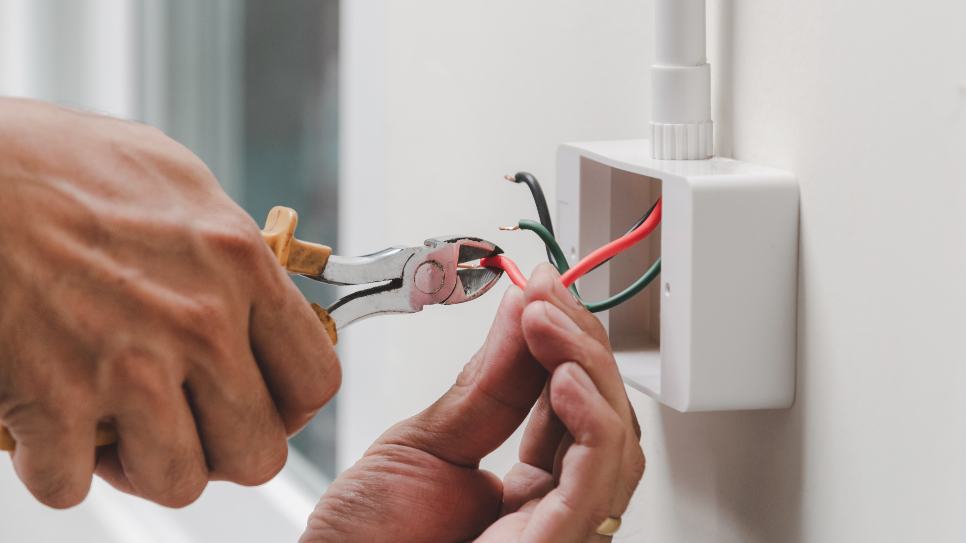 Hands use pliers to wire a white electrical outlet box with red and black cables.