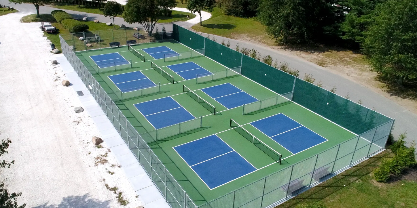 Aerial view of several pickleball courts, blue playing surfaces and green surrounding areas, enclosed by a fence. Aerial view of several pickleball courts, blue playing surfaces and green surrounding areas, enclosed by a fence.