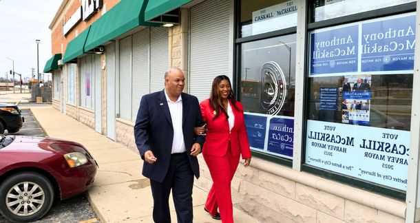 Two people walking outside a storefront with blue window signs and a parked maroon car