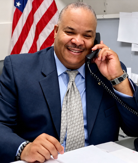 Smiling man in a navy suit talking on a phone at a desk with an American flag in the background