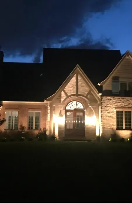 Well-lit stone house at dusk with a peaked entry and dark roof under a blue evening sky