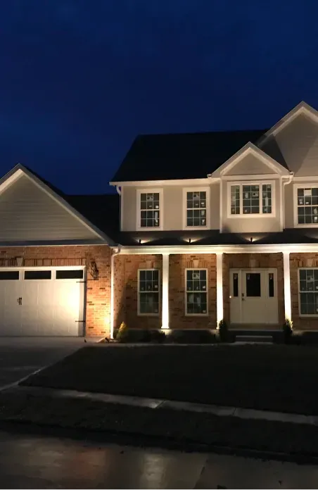 Two-story suburban house at dusk with porch lights on and a lit garage against a dark blue sky