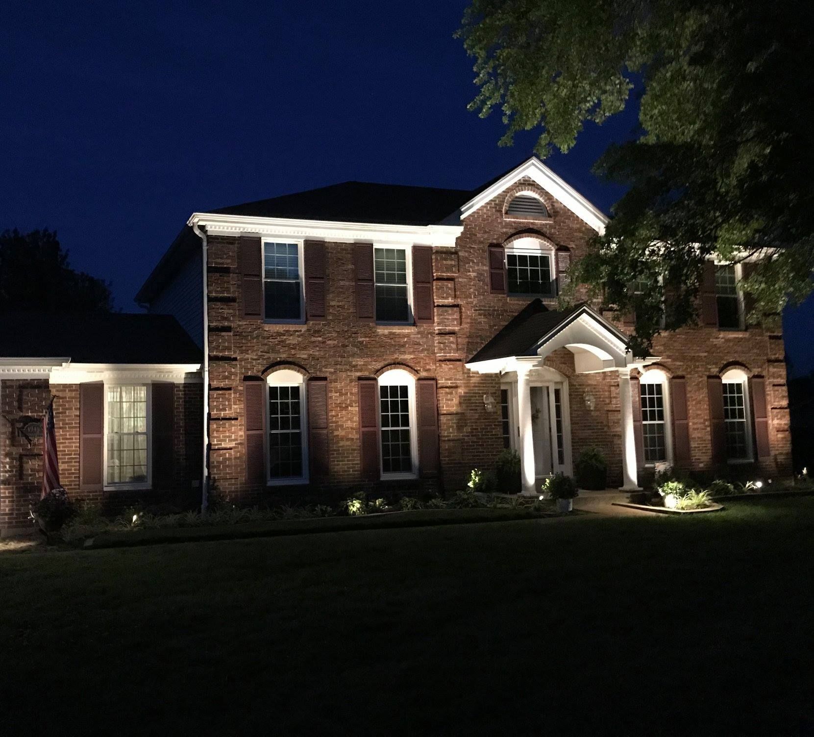 Brick house at night with lit windows, front porch, and landscaped lawn under a dark blue sky
