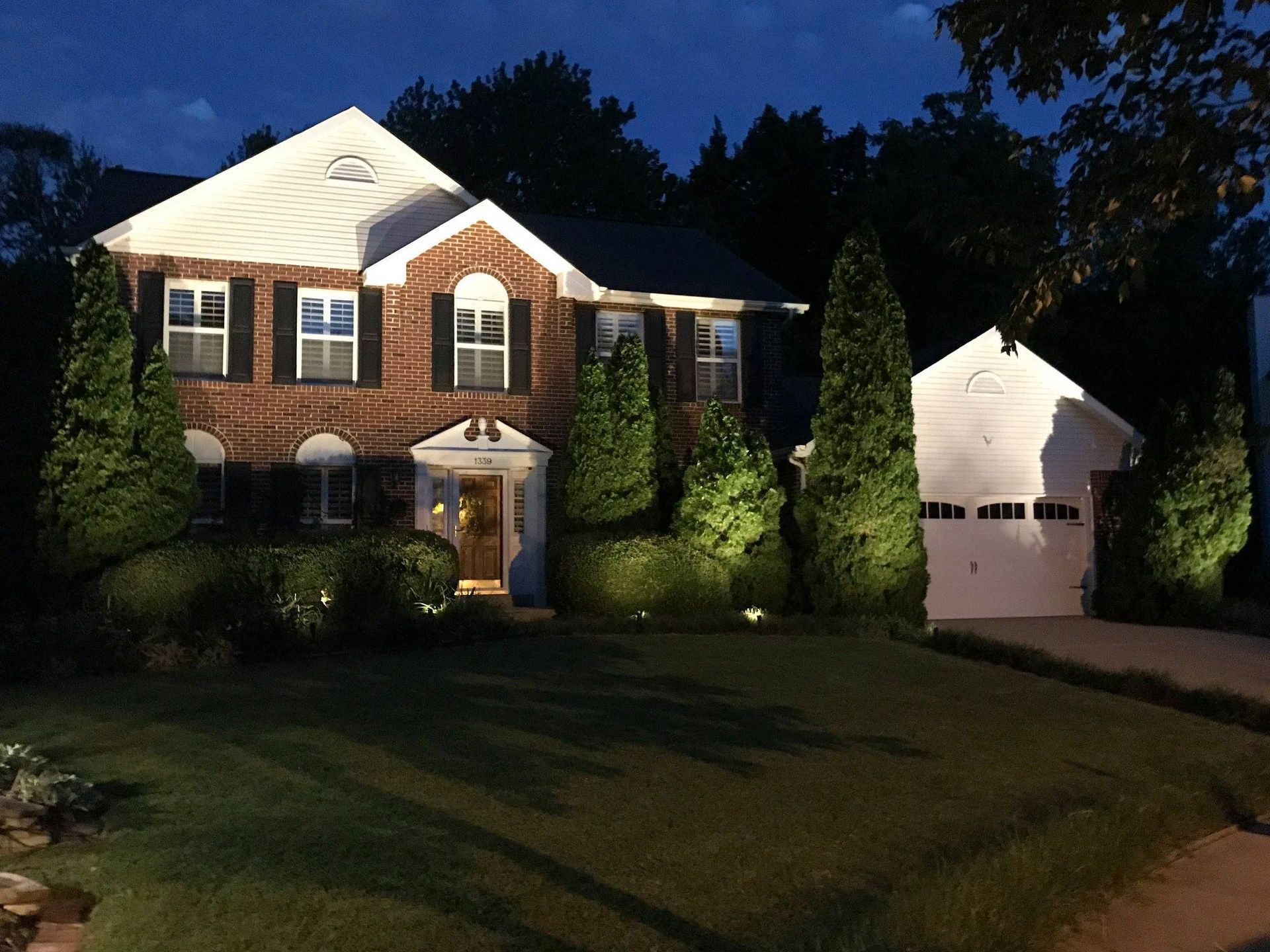Brick house with lit windows and a driveway at dusk, with a detached garage on the right.