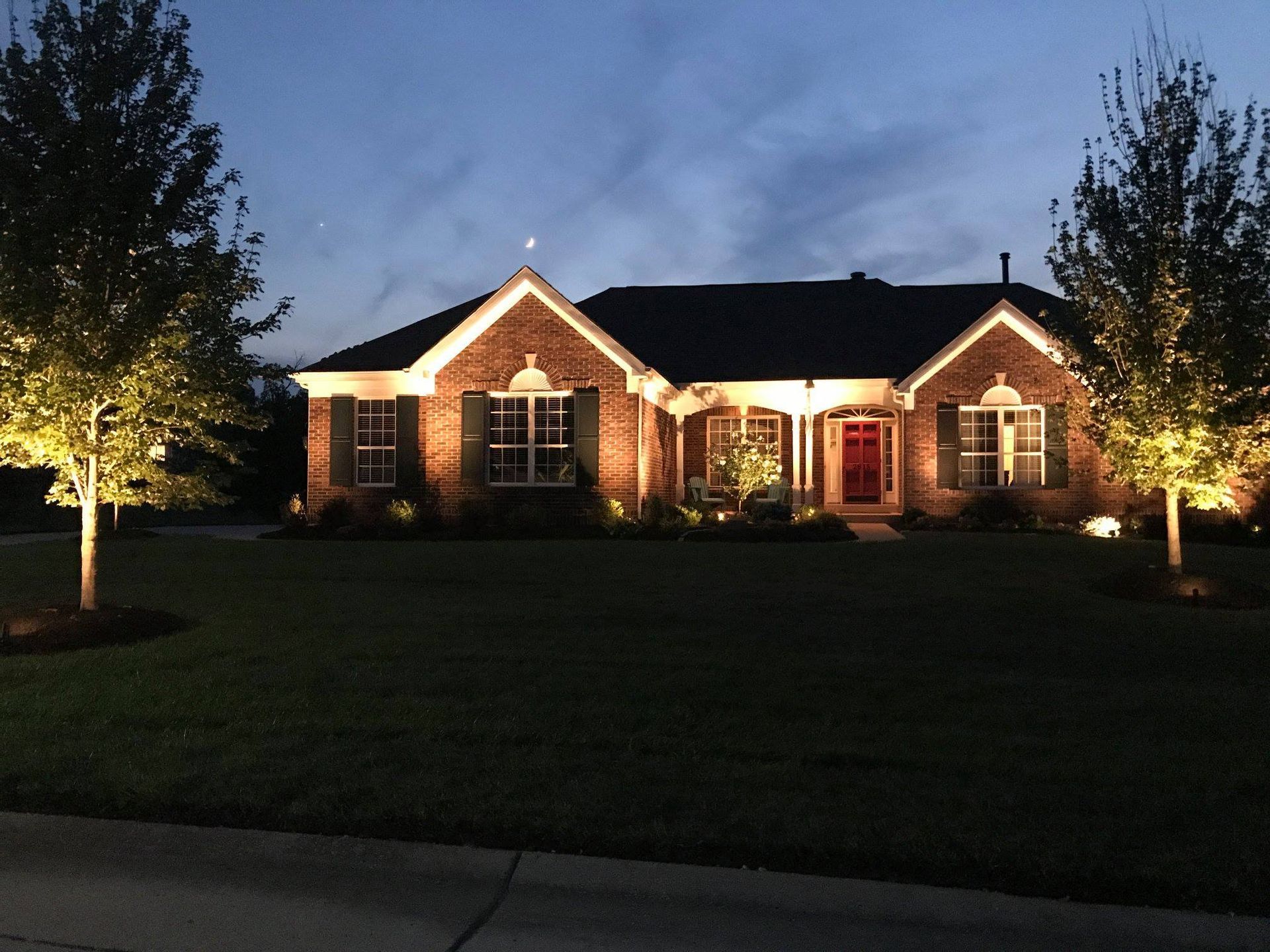 Brick house lit up at dusk with warm landscape lights and trees framing the front yard