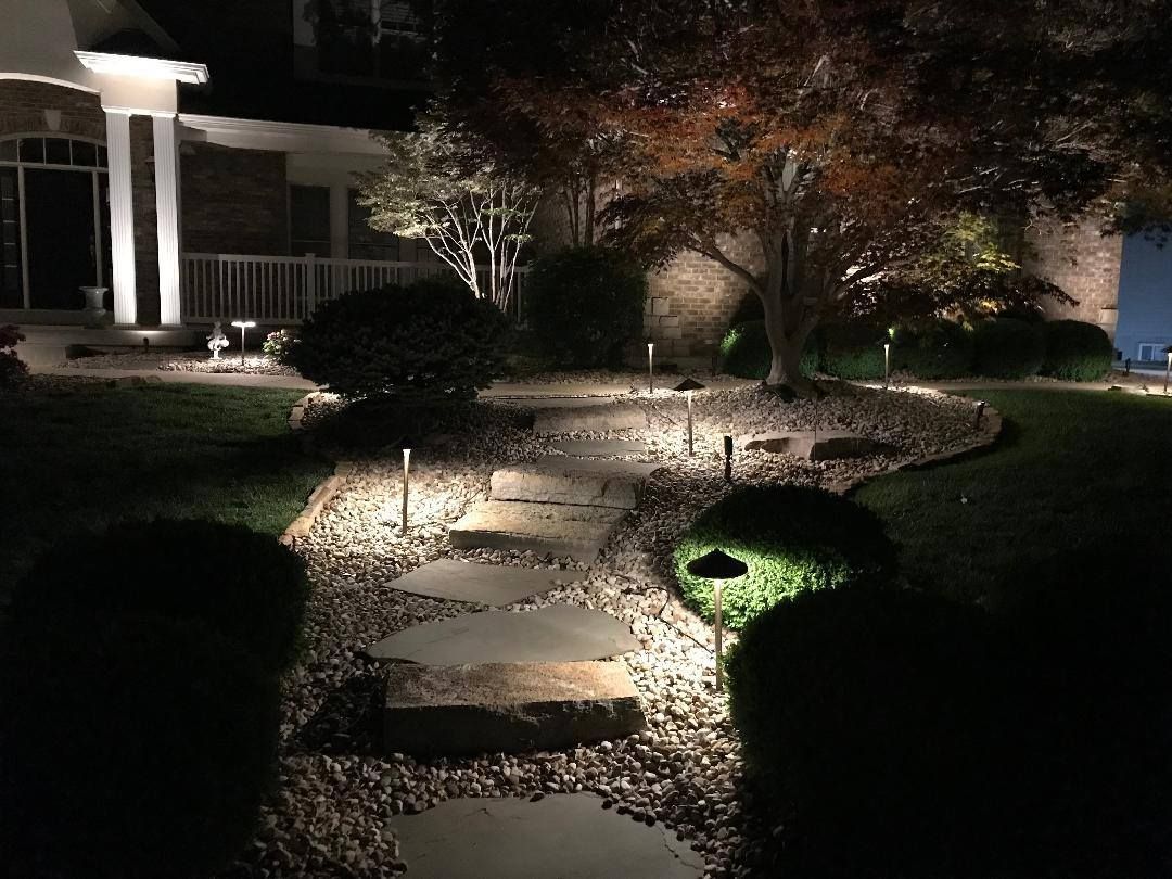 Lit stone walkway through a landscaped garden at night, illuminated by ground lights beside a house.