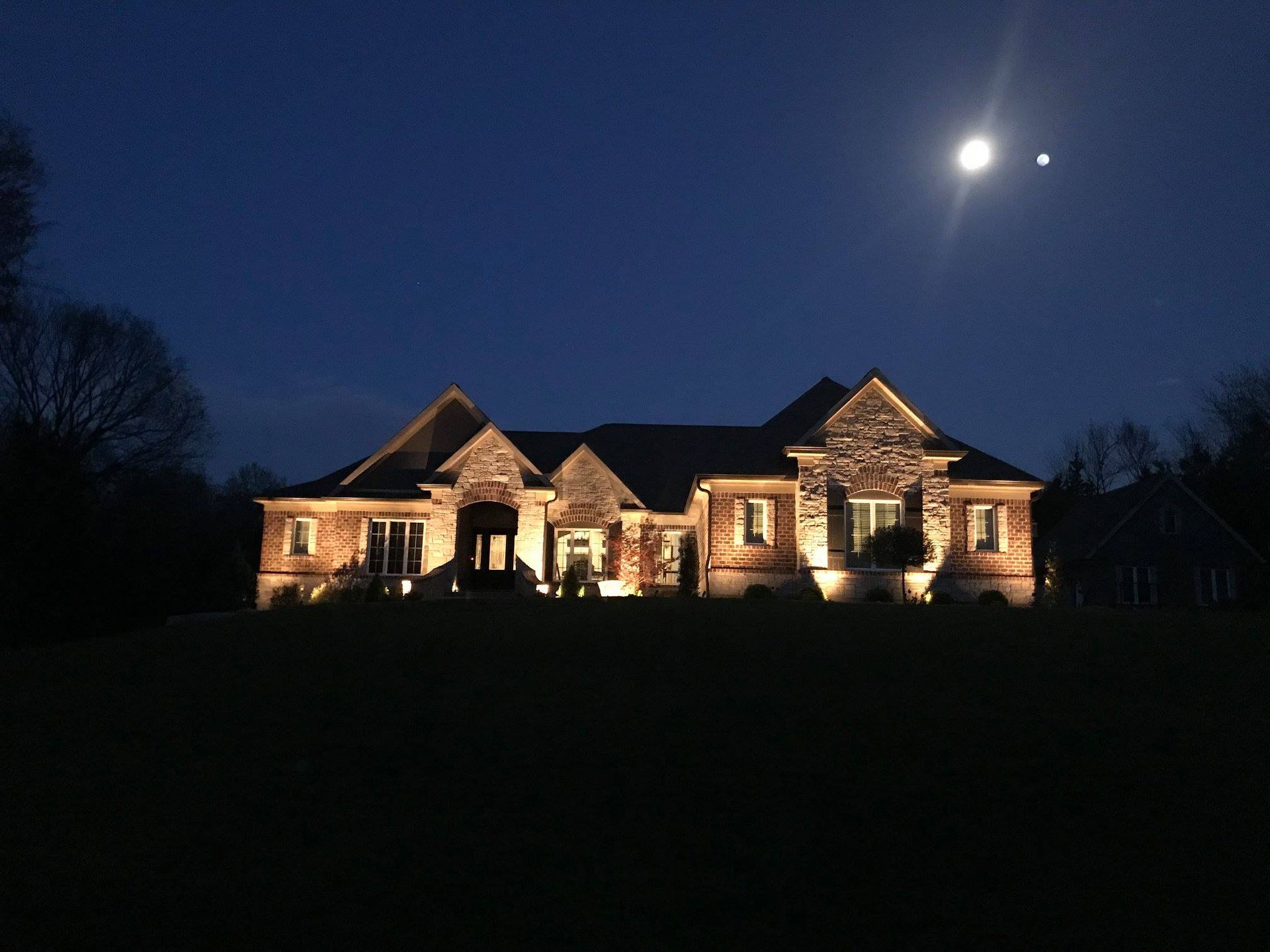 Well-lit stone house at night under a dark blue sky with the moon overhead