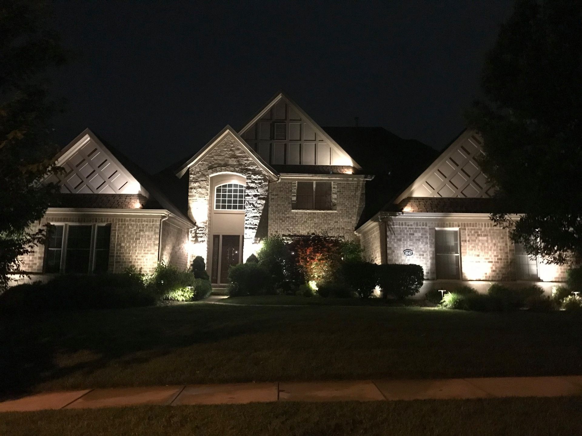 Illuminated stone house at night with peaked roofs and landscaped front yard